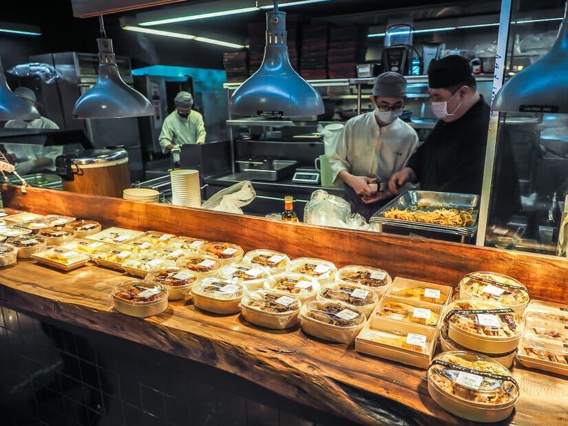 A wooden bar with some take-away meals on it and some chefs in the kitchen behind it