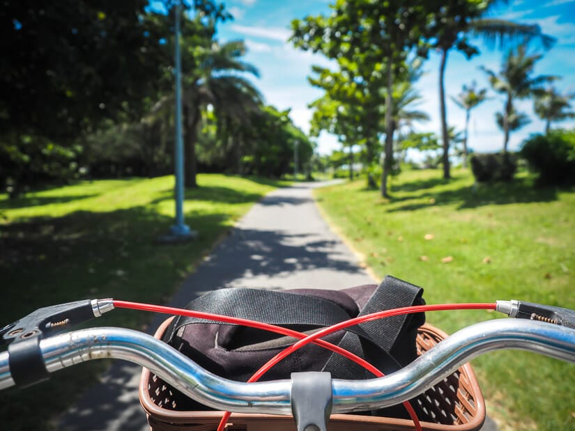 The handlebars of a bike, with a black camera bag in the basket, with the Cijin Coastal Park bike path in front