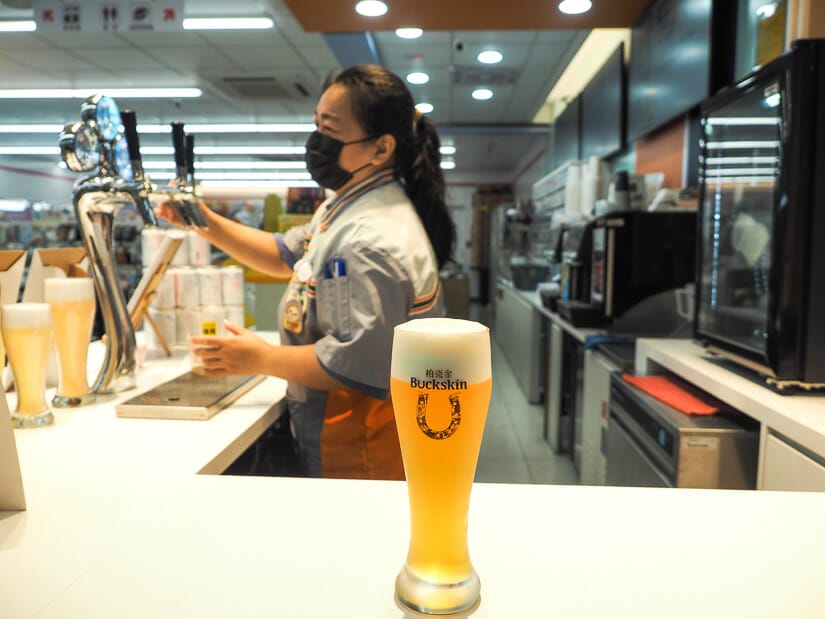 A glass of draft beer on a counter with a 7-11 employee behind it pouring another one in Liuhe Night Market