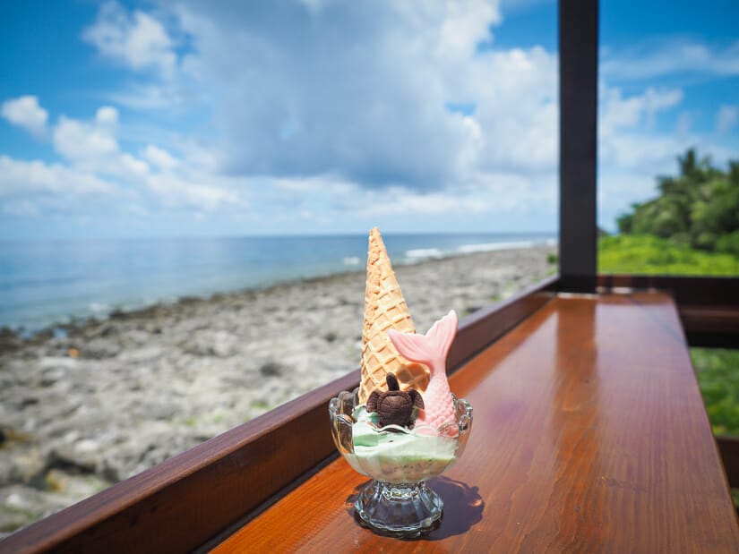 A wooden bar with a cup of ice cream on it with a mermaid tail, chocolate sea turtle, and the coast of Xiaoliuqiu in the background