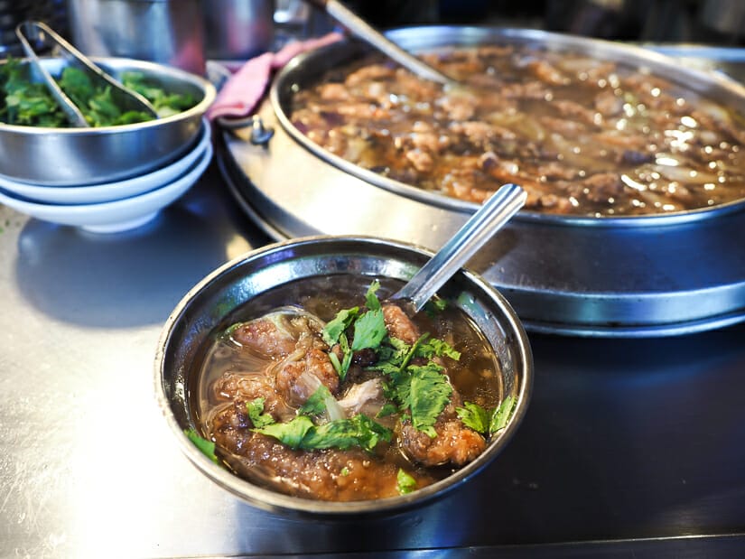 Close up of a bowl of braised eel soup with a big vat of the soup in the background 