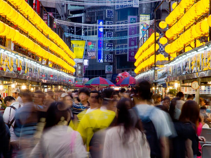 Crowds of people in Keelung Night Market, made fuzzy by their movement, and rows of yellow lanterns lining either side
