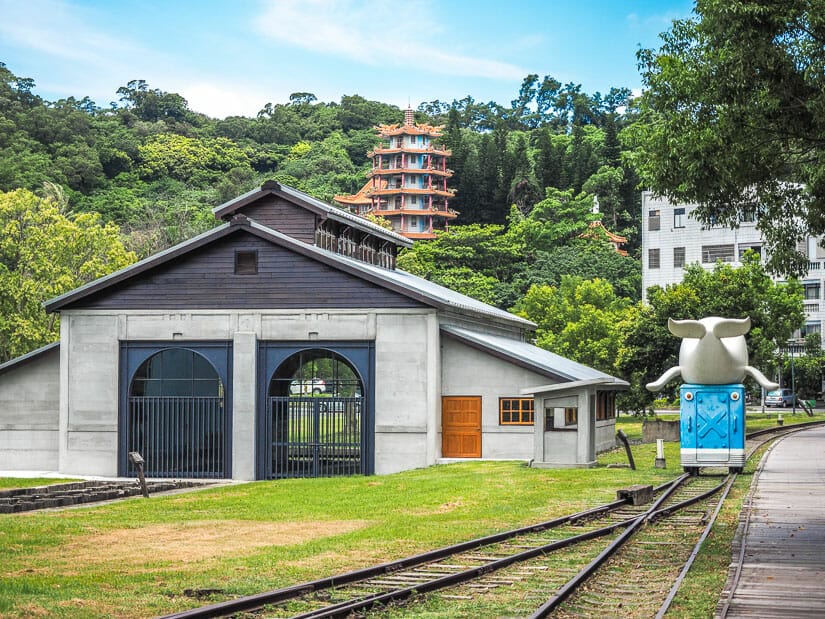 A railway line leading to a train warehouse at Taitung Railway Art Village, with a cute whale statue and wooden walking path on the right, and a pagoda on a forested hill in the background