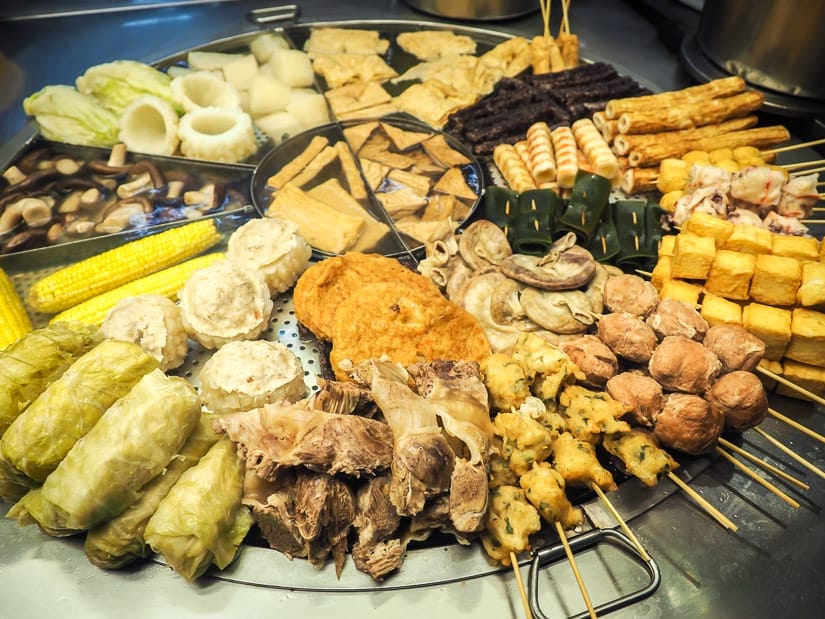 A massive circular metal pot building into a metal counter, full of dozens of sticks of various meats, tofus, fish cakes, corn on the cobs, and more, all stewing in the soup