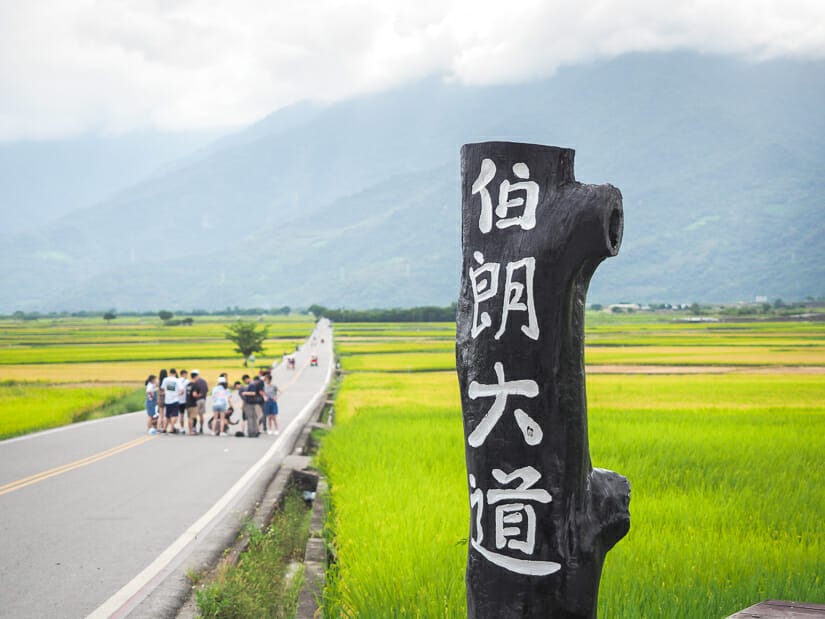 A group of people standing on the road on Mr. Brown Avenue in Chishang, with a pole in the foreground that says Brown Boulevard in Mandarin characters