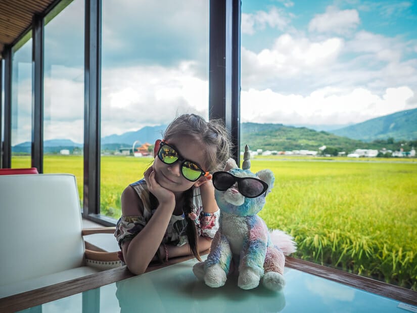 A girl and her teddy unicorn, both wearing sunglasses, in a cafe in Chishang with rice paddies outside the window
