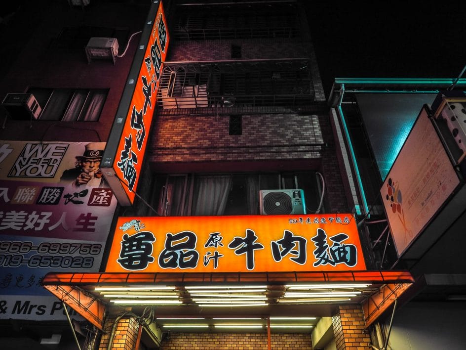 Looking up at a horizontal and a tall vertical orange and black sign of a beef noodle shop in a night market