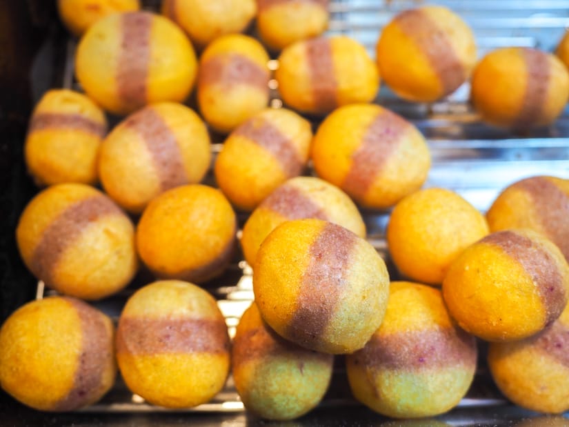 Close up of a pile of tri-color (orange, purple, and yellow) deep fried sweet potato balls cooling on a metal rack