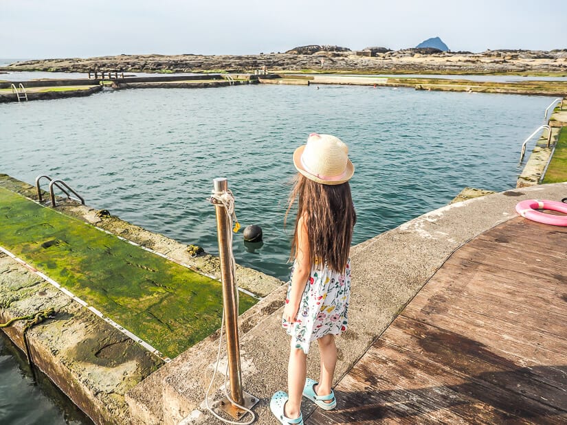 A young girl standing beside a natural seawater pool, looking down at it.