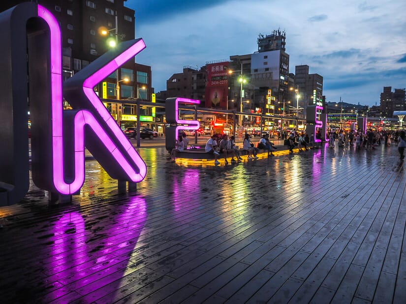 Giant neon purple letters that spell KEELUNG on a wooden boardwalk at Keelung Martime Plaza, with some people sitting on benches