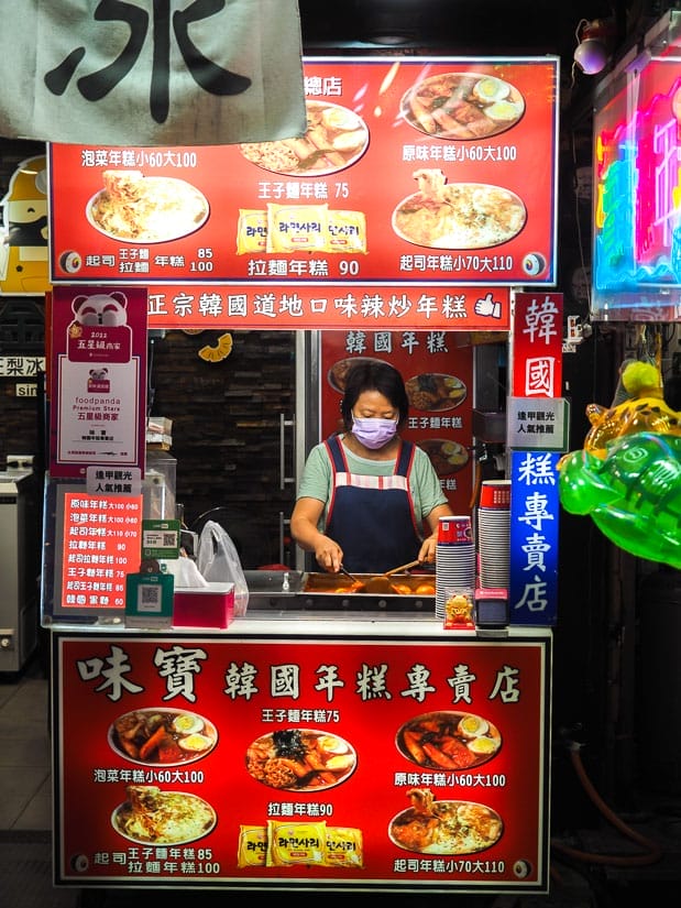 Vertical image of a food stall in a night market, with red signage on bottom and top, a woman wearing mask and preparing food behind it, and menu with pictures