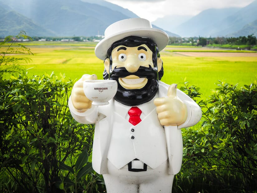 A statue of Mr. Brown, the mascot of a Taiwanese coffee company, standing in front of rice paddies on Brown Boulevard