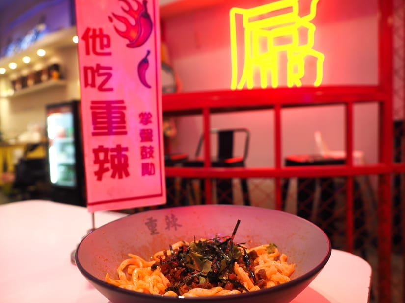 A bowl of noodles on a table, with a sign beside it with chili pepper symbols and Mandarin characters which say "he's eating super spicy", and a yellow neon light on the wall behind, and the whole photo has a red glow to it