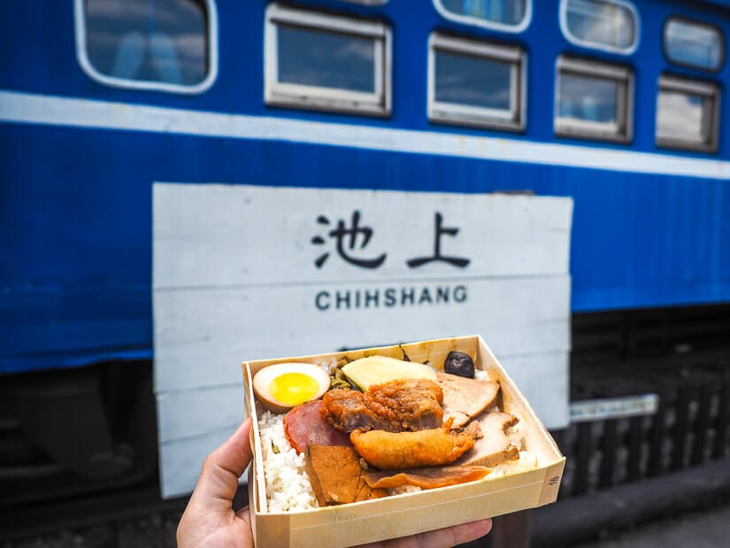 A hand holding up one of the famous lunchboxes from Chishang in front of an old blue train at Wutao Chishang Lunchbox Museum