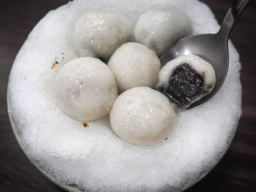 A bowl of shaved ice with 6 tang yuan balls on top. One is split open with black sesame sauce spilling out and a spoon is lifting it up