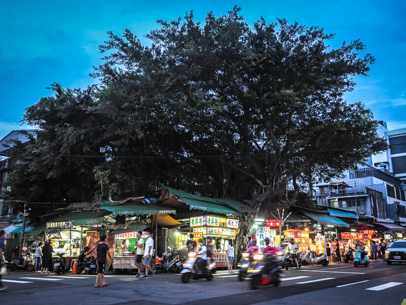 Scooters drive by under a tree at Zhongxiao Night Market in Taichung, with food vendors along the road under the tree