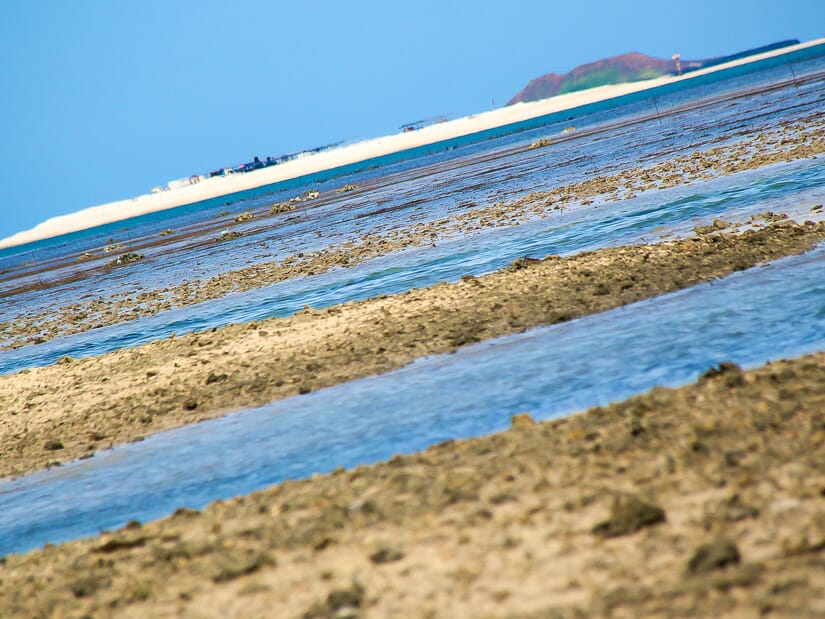 A thing strip of white sand visible in the distance across some shallow water