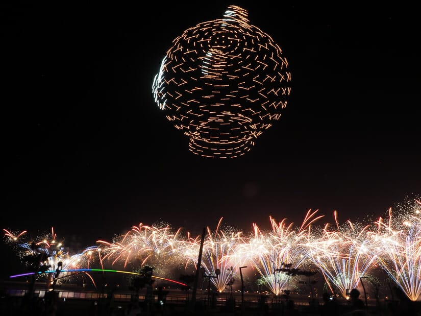 Fireworks shooting off and lit up drones in a black sky lit up in the shape of a bear's head, with a bridge with rainbow colored lights below at the Penghu International Fireworks Festival