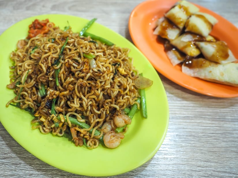 Two plates of food on a table. Left side is a green plate with fried instant noodles and shrimps. Left side plate or orange and has a green onion cake doused in thick soy sauce