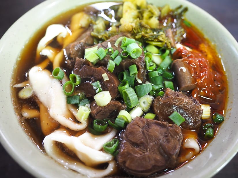 Close up of a bowl of beef noodles, with chunks of beef, wide white noodles, spicy sauce, pickled greens, and green onions as garnish