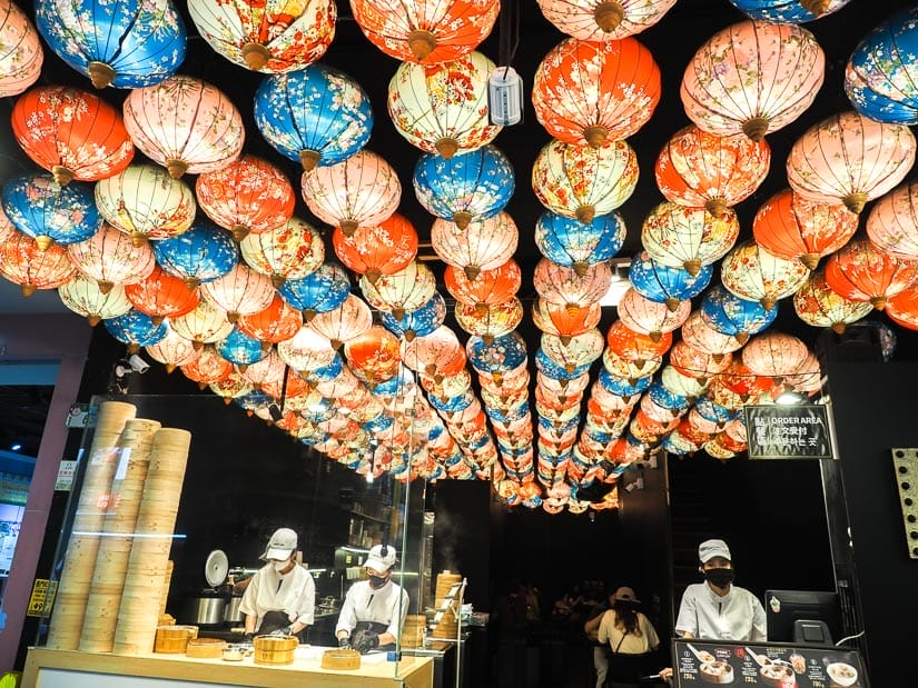 Looking into a restaurant with long rows of many colorful Chinese lanterns hanging from the ceiling and some cooks at work behind a counter with baskets of steaming Xiao Long Bao