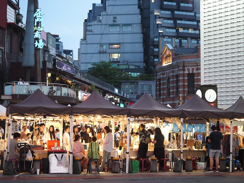 An outdoor craft market in the early evening, with many tents with lights and products on display and people shopping for them