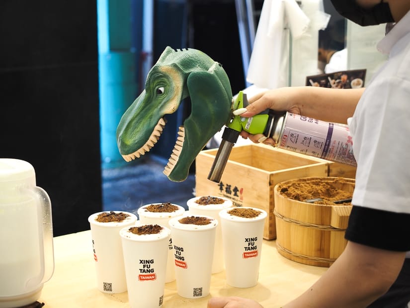 A hand holding a torch which has a dinosaur head on the top and using it to burn the top of some bubble tea drinks