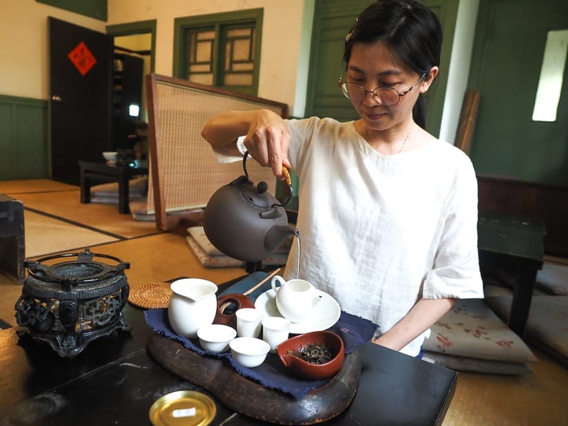A woman kneeling beside a low table and pour water from a kettle into a tea pot