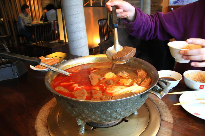 A hot pot with three legs, spicy soup, and someone is scooping some out