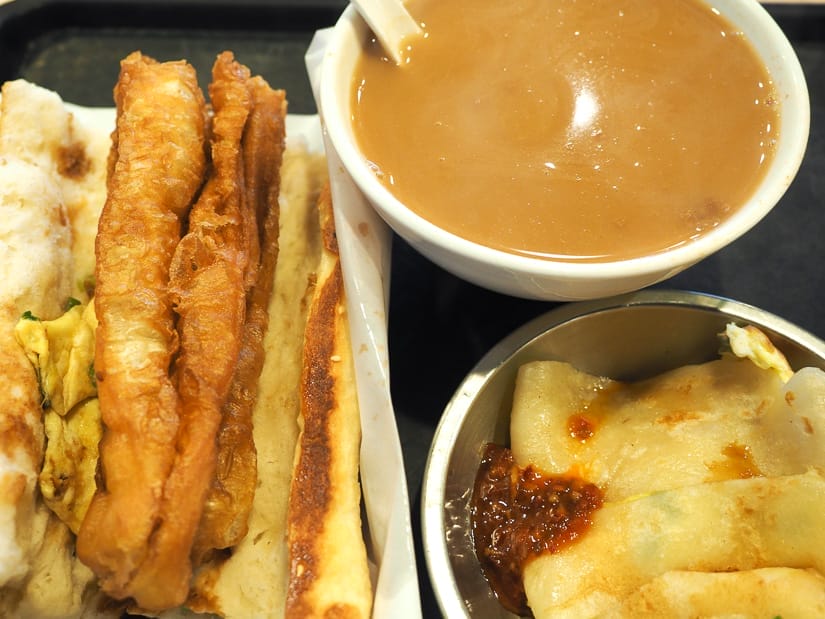 Three Taiwanese breakfast items on a table – baked roll with fried dough stick, egg crepe in a metal bowl with spicy sauce, and bowl of brown rice milk with spoon in it