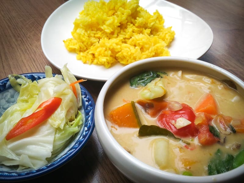 Three dishes on a table: one thai curry with lots of vegetables, one yellow rice, and one pickled vegetables