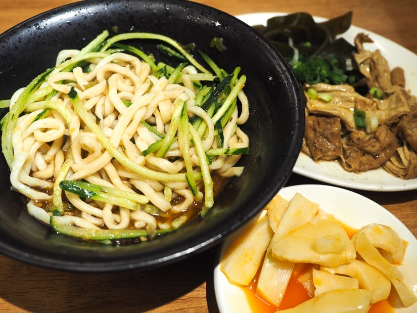 A large black bowl of noodles with cucumber strips with two vegetable side dishes