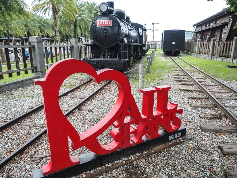 A black locomotive on railway tracks with red sign in the foreground that says I heart rail fans