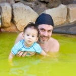 Nick Kembel holding his young son while bathing in a bright yellow hot spring tub, both wearing swimming caps