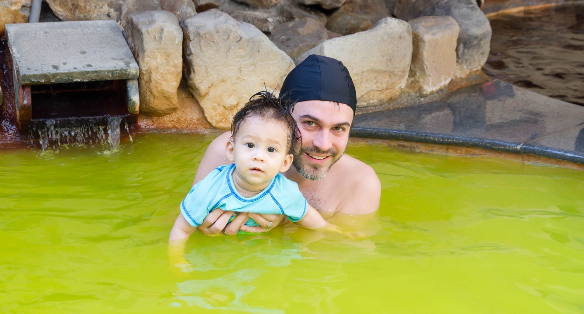 Nick Kembel holding his young son while bathing in a bright yellow hot spring tub, both wearing swimming caps