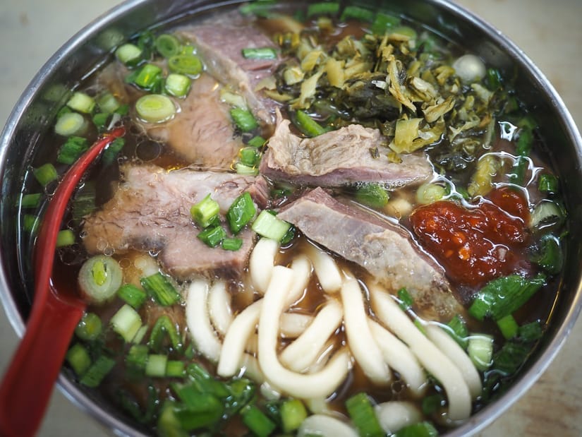 Close up of a metal bowl of soup with noodles, chunks of beef, and green onions