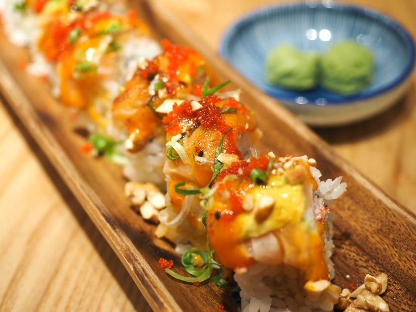 Close up of a long wooden tray of sushi rolls dressed with many sauces and garnishes and small bowl of wasabi to the side