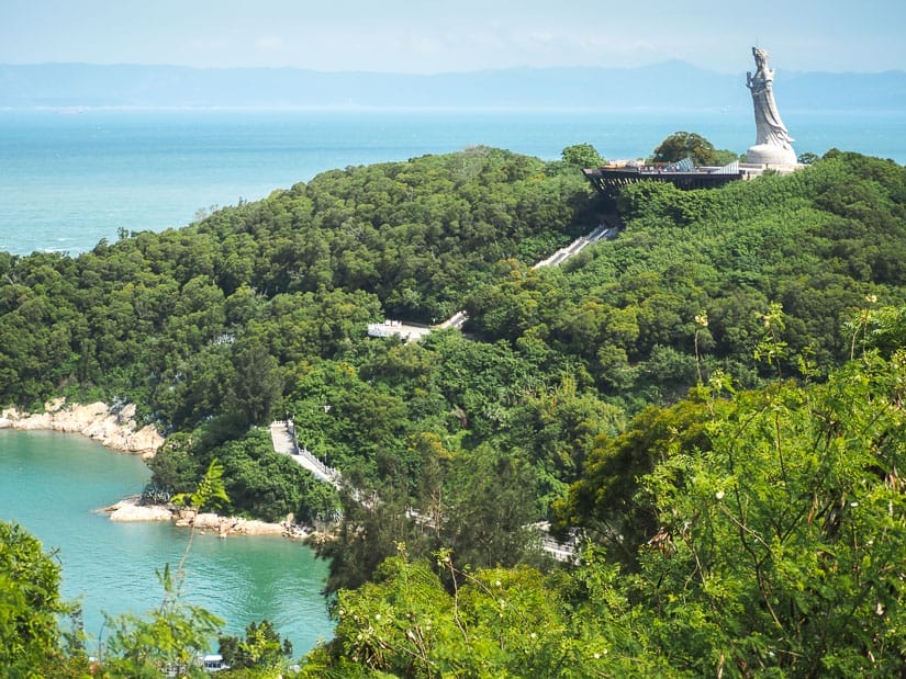 A tall statue of Matsu on a tree covered hill overlooking the sea with long set of stair cases going from the beach all the way up to it
