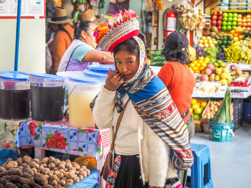An indigenous Quechua woman with colorful clothing and hat looks at the camera while shopping in a traditional market in Ollantaytambo