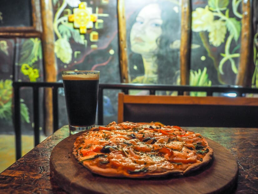 A pizza and dark beer on a table with painting of a woman's face on the wall behind
