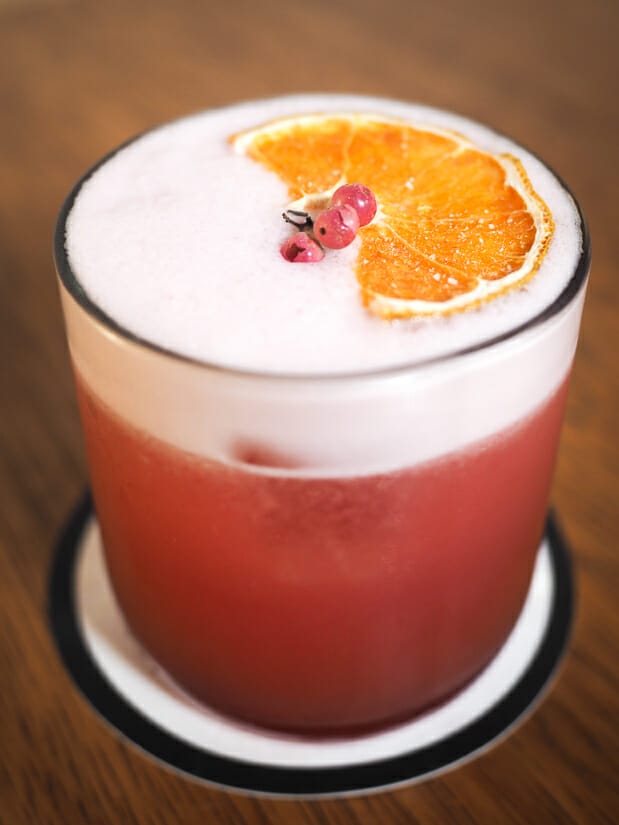 Close up of a pink cocktail with orange slice and little pink berries as garnish