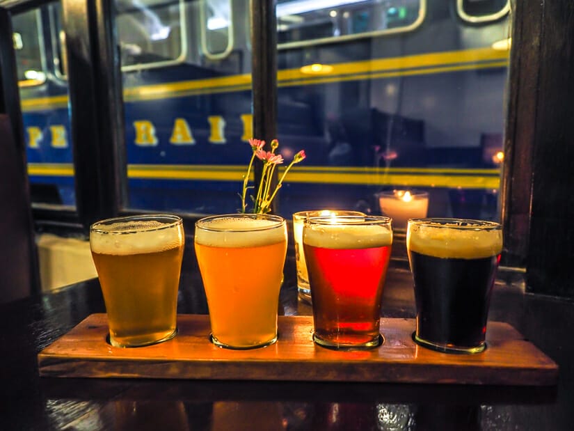 A row of beer samples with candle and flower behind them, and large window with blue Inca Rail train passing by