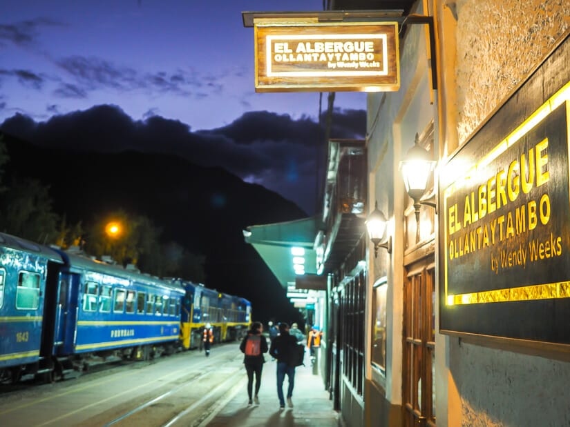 Exterior signs of El Albergue restaurant, with people walking along the platform of Ollantaytambo train station