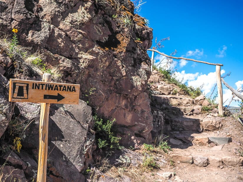 Sign that says "Intihuatana" and points to set of stairs going up behind a large rock