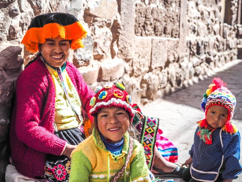 A Quechua woman and her two children wearing colorful clothing and sitting against a wall in Pisac