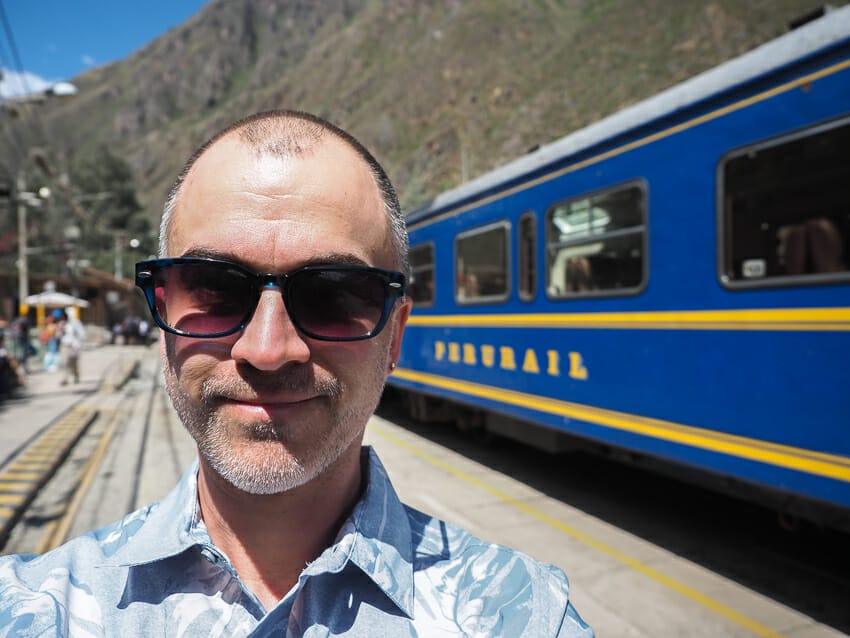 A selfie of me with a blue Peru Rail train behind me, on its way to Machu Picchu