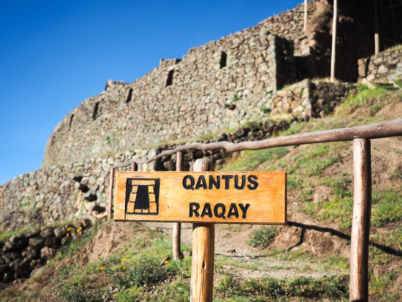 A sign that says Qantus Raqay with Incan ruins behind it at Pisac
