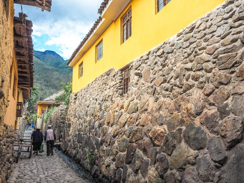 Two Quechu people walking down a narrow cobblestone alleyway in the Qosqo Ayllu of Ollantaytambo, with a stone and yellow building forming one wall