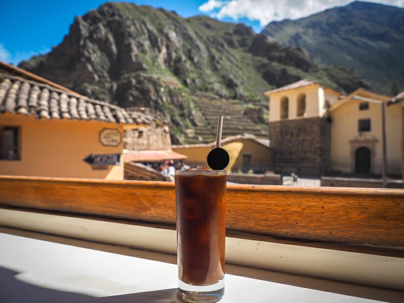 A glass of iced coffee on a wooden counter with a church and Ollantaytambo ruins in the background
