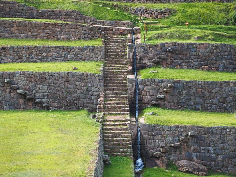 Ruins of an Inca stone staircase with grassy terraces to the sides and water canal flowing down beside the stairs
